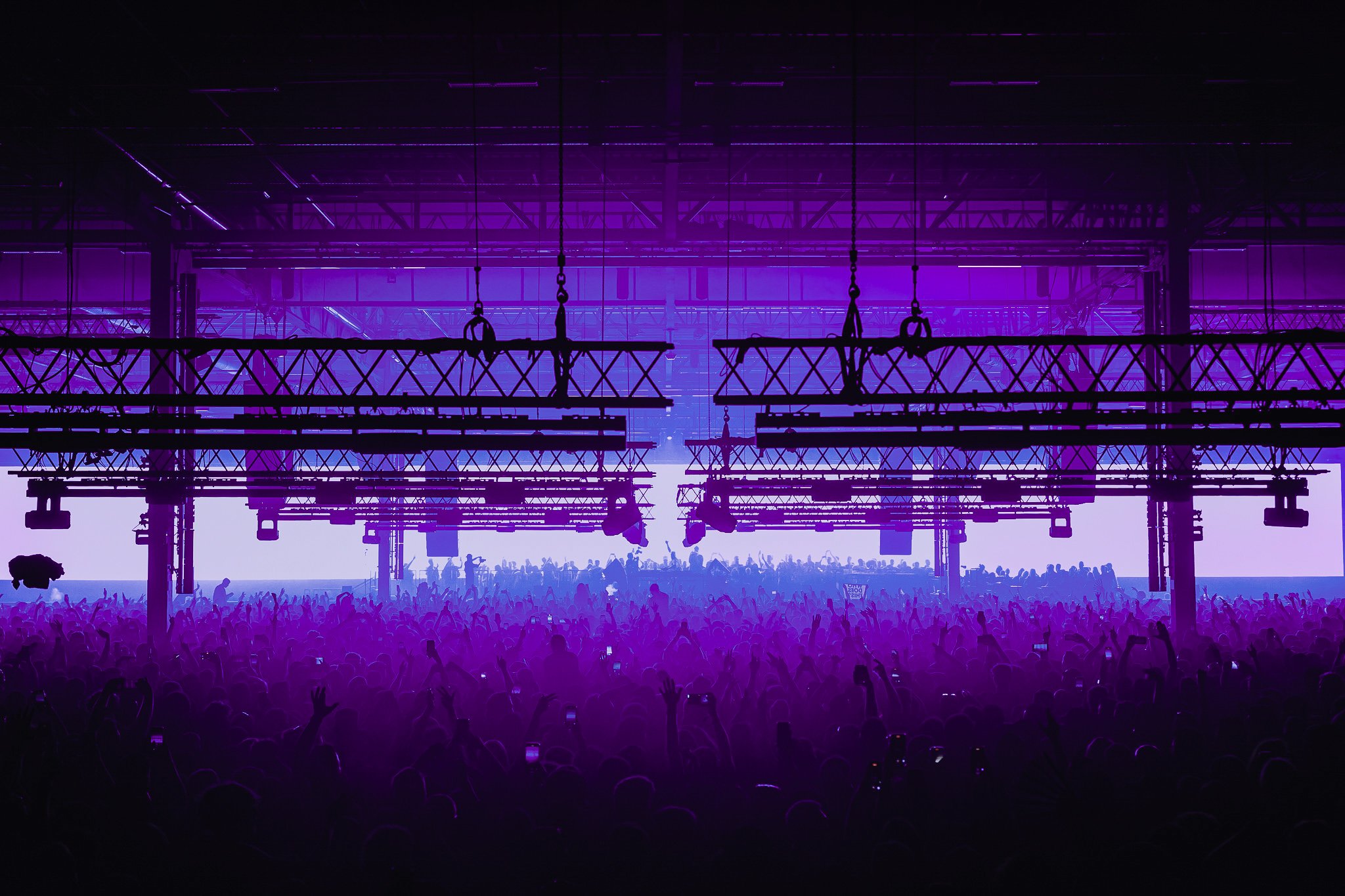A large indoor concert or rave with a dense crowd beneath hanging lighting rigs, illuminated by vibrant purple and blue stage lights.